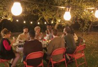 Друзья наслаждаются ужином в саду под деревьями с волшебными огнями — стоковое фото