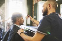 Male barber spraying hair of man in barbershop — Stock Photo