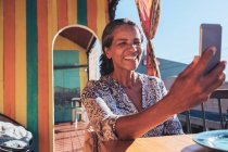 Усміхаючись, щаслива жінка бере селфі зі смартфоном на сонячному патіо — стокове фото