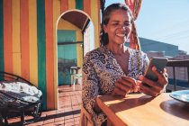 Souriant, femme heureuse en utilisant smartphone sur balcon restaurant ensoleillé — Photo de stock