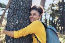 Fröhliche, unbeschwerte junge Wanderin umarmt Baum im sonnigen Wald — Stockfoto