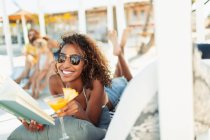 Porträt einer glücklichen, unbeschwerten jungen Frau, die Buch liest und am Strand einen Cocktail trinkt — Stockfoto