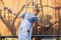 Travailleur masculin sur échelle tachant le parement de bois sur l'extérieur de la maison — Photo de stock