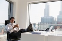 Empresário sorridente usando telefone inteligente com os pés na mesa no escritório moderno, ensolarado e urbano — Fotografia de Stock