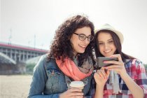 Jóvenes amigas usando teléfono inteligente y tomando café al aire libre - foto de stock