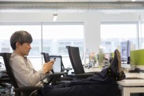 Empresario usando teléfono inteligente con los pies en el escritorio de la oficina - foto de stock