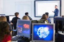 Молодші студенти за комп'ютерами, які спостерігають за вчителем на проекційному екрані в класі — стокове фото