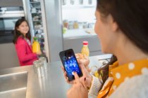 Mãe usando o aplicativo de telefone inteligente para rastrear mantimentos na geladeira, assistindo filha — Fotografia de Stock