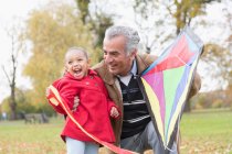 Playful grandfather and granddaughter flying a kite in autumn park — Stock Photo