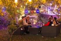 Friends talking and drinking under trees with string lights at garden party — Stock Photo