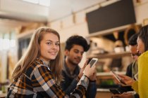 Retrato confiado joven mujer usando el teléfono inteligente en la cafetería con amigos - foto de stock
