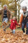 Бабушка с дедушкой и внучка пинают осенние листья в парке — стоковое фото
