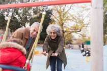 Играющие бабушка и дедушка толкают внука на качели на детской площадке — стоковое фото