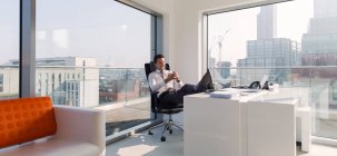 Uomo d'affari con i piedi sulla scrivania in ufficio soleggiato, moderno, urbano — Foto stock
