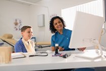 Female pediatrician and boy patient talking, using computer in doctors office — Stock Photo