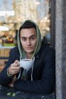 Retrato confiante jovem em cappuccino bebendo com capuz — Fotografia de Stock