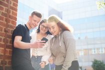 Teenage friends using smart phone outside sunny school building — Stock Photo