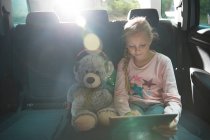Menina com ursinho de pelúcia usando tablet digital no banco de trás do carro — Fotografia de Stock