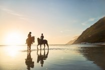 Mujeres jóvenes a caballo en el océano tranquilo surf al atardecer - foto de stock