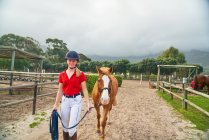 Portrait confiant adolescent fille leader cheval le long des paddocks ruraux — Photo de stock