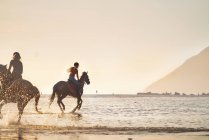 Young women horseback riding in ocean surf at sunset — Stock Photo