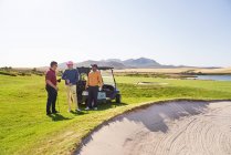Hombres amigos golfistas hablando en la trampa de arena en el campo de golf soleado - foto de stock