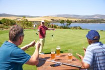 Felices golfistas varones bebiendo cerveza y practicando poner en el campo de golf - foto de stock