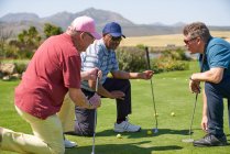 Los golfistas masculinos de rodillas y hablando en la práctica soleada de poner verde - foto de stock