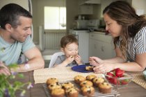 Familia joven comiendo magdalenas frescas en la cocina - foto de stock