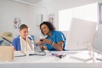 Pediatra feminina que prescreve medicação para menino com braço na funda em consultório médico — Fotografia de Stock