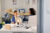 Жінка-педіатр розмовляє з хлопчиком пацієнткою з рукою в слінгу в офісі лікарів — стокове фото