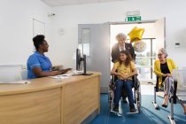 Woman pushing girl patient in wheelchair into clinic reception — Stock Photo
