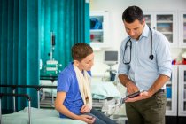 Pediatra masculino mostrando tableta digital a niño paciente con brazo en honda en el hospital - foto de stock