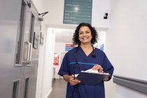 Retrato sonriente, confiada doctora con historial médico, haciendo rondas en el pasillo del hospital - foto de stock