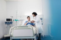 Médecin avec tablette numérique faisant des rondes, parlant avec le patient se reposant dans le lit d'hôpital — Photo de stock