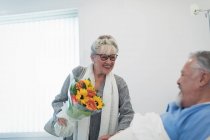 Glückliche Seniorin bringt Blumenstrauß zu Mann, der sich im Krankenhauszimmer erholt — Stockfoto