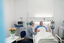 Старша жінка, яка відвідує, заспокоює чоловіка, який відпочиває в лікарняному ліжку — стокове фото