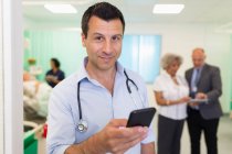 Retrato confiado médico masculino usando el teléfono inteligente en la sala de hospital - foto de stock