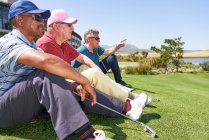 Golfistas masculinos descansando sentados na grama no campo de golfe ensolarado — Fotografia de Stock