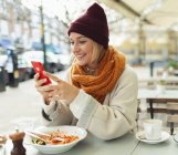 Sorrindo jovem com telefone inteligente comendo almoço no café calçada outono — Fotografia de Stock