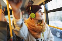 Mujer joven en gorra de siembra y bufanda escuchando música con auriculares en autobús. - foto de stock
