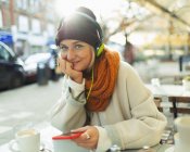 Retrato mujer joven confiada escuchando música con audífonos y reproductor de mp3 en la cafetería sidewalk de otoño. - foto de stock