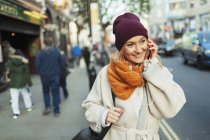 Молода жінка в панчішній шапці і шарфі говорить на смартфоні на міському тротуарі — стокове фото