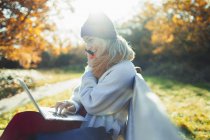 Mulher nova usando laptop e falando em telefone inteligente no banco ensolarado do parque de outono — Fotografia de Stock