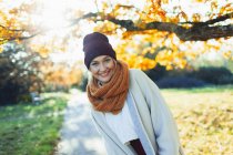 Portrait happy young woman in stocking cap and scarf in sunny autumn park — Stock Photo