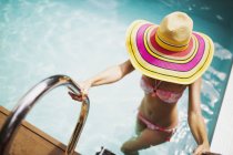 Mujer con sombrero de sol y bikini saliendo de la soleada piscina de verano - foto de stock