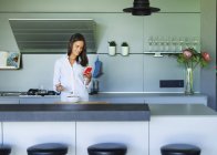 Mujer usando el teléfono inteligente y desayunando en la cocina moderna - foto de stock