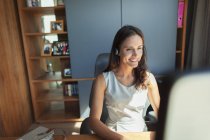 Усміхнена бізнес-леді з гарнітурою, що працює за комп'ютером в домашньому офісі — стокове фото