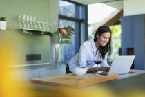 Smiling woman using laptop and drinking red wine in kitchen — Stock Photo