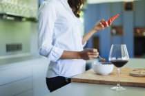 Mujer comiendo ensalada y bebiendo vino tinto, usando un teléfono inteligente en la cocina - foto de stock